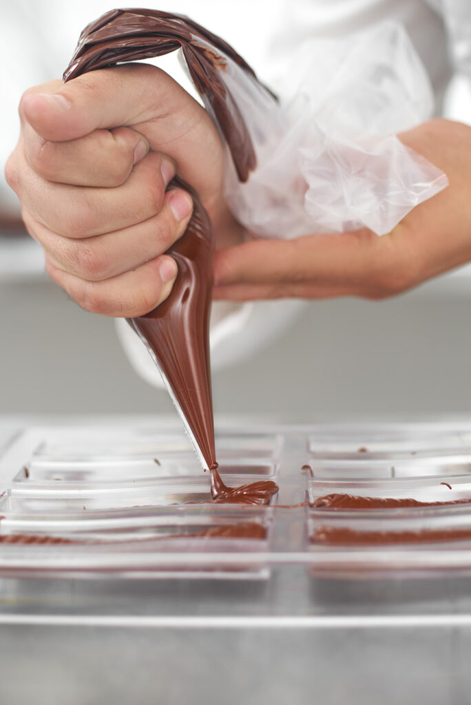 production, food preparation and people concept - confectioner with piping bag squeezing liquid chocolate into chocolate bar shape in pastry shop